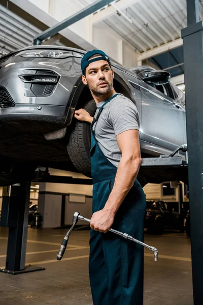 Young auto mechanic with lug wrench standing at car at mechanic shop — Stock Photo