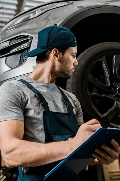 Reparador concentrado en uniforme con el bloc de notas comprobar las ruedas del coche en el taller de reparación de automóviles - foto de stock