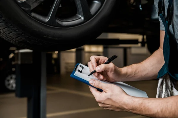 Recortado tiro de reparador con bloc de notas de control de ruedas de coche en taller de reparación de automóviles - foto de stock