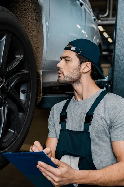 Side view of concentrated repairman with notepad checking car wheels at auto repair shop — Stock Photo