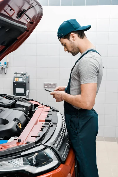 Auto mechanic in uniform using smartphone at car with opened cowl at mechanic shop — Stock Photo