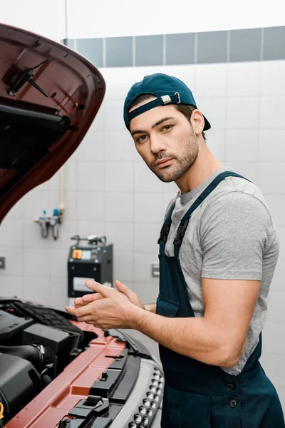 Side view of repairman in uniform standing at car with opened cowl at auto repair shop — Stock Photo