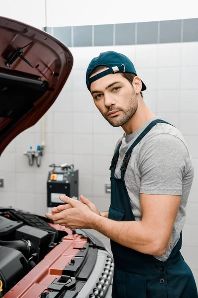 Vista lateral del reparador en uniforme de pie en el coche con capucha abierta en el taller de reparación de automóviles — Stock Photo