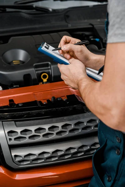 Vista parcial del reparador con bloc de notas que examina el coche en el taller de reparación de automóviles - foto de stock