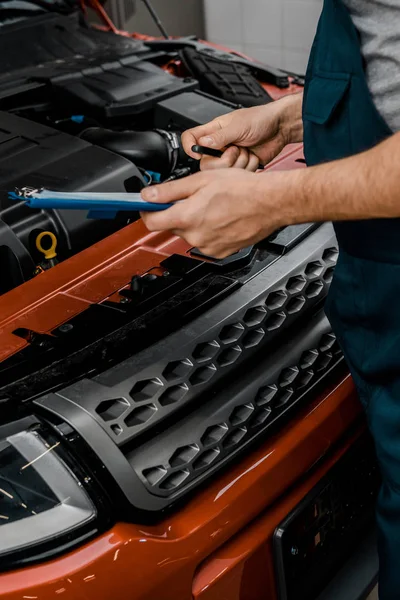 Teilansicht eines Werkstattmitarbeiters mit Notizblock, der das Auto in der Autowerkstatt untersucht — Stockfoto