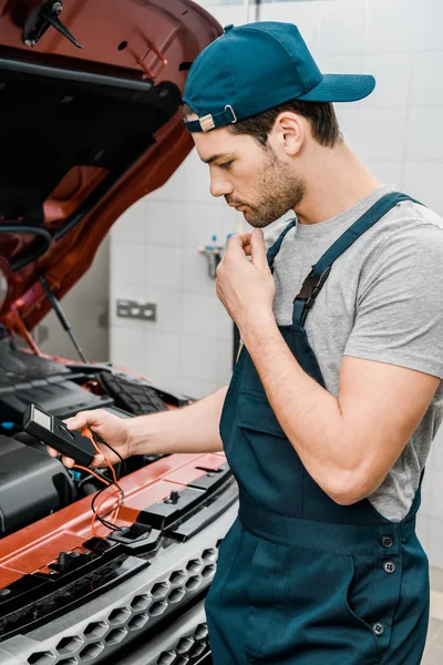 Vista laterale del meccanico automatico con il voltmetro multimetro che controlla la tensione della batteria dell'automobile al negozio meccanico — Foto stock