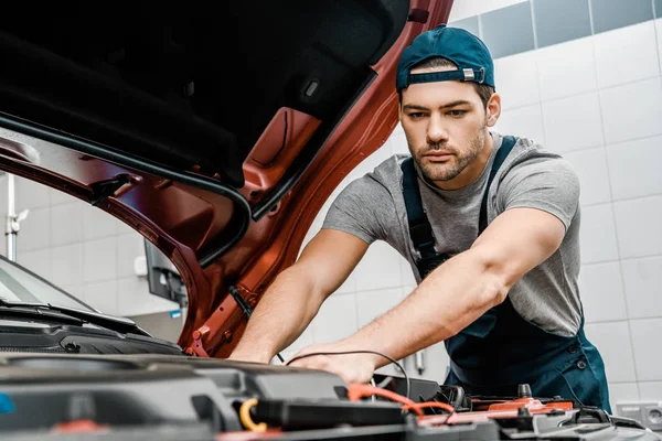 Retrato de joven mecánico de automóviles con voltímetro multímetro comprobación de voltaje de la batería del coche en el taller mecánico — Stock Photo