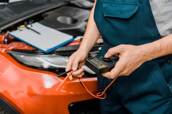 Tiro recortado de auto mecânico segurando multímetro voltímetro para verificação de tensão da bateria do carro na loja mecânica — Fotografia de Stock