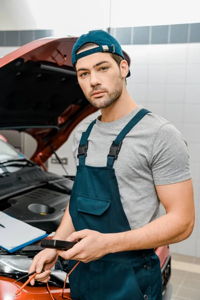 Porträt des Automechanikers, der ein Multimeter-Voltmeter für die Spannungskontrolle der Autobatterie in der Werkstatt hält — Stockfoto
