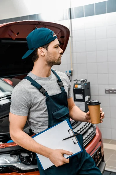 Nachdenklicher Mechaniker mit Coffee to go und Notizblock an Auto mit geöffneter Motorhaube gelehnt — Stockfoto