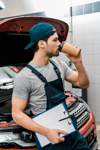 Joven mecánico con bloc de notas bebiendo café y apoyado en el coche con capucha abierta en el taller mecánico - foto de stock