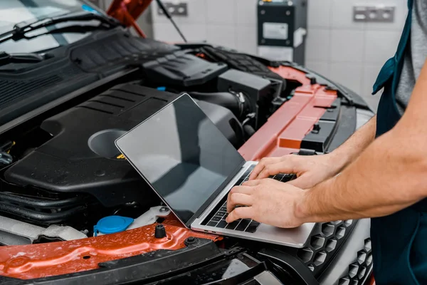 Tiro recortado de mecánico de automóviles que trabaja en el ordenador portátil con pantalla en blanco en el automóvil con la capucha de coche abierto en el taller mecánico - foto de stock