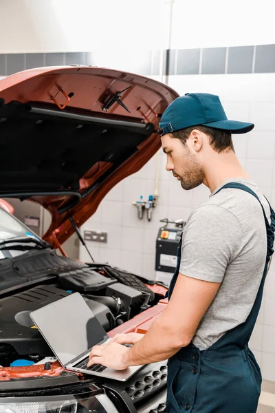 Vista lateral del mecánico automático que trabaja en el ordenador portátil en el automóvil con la capucha abierta del coche en el taller mecánico — Stock Photo