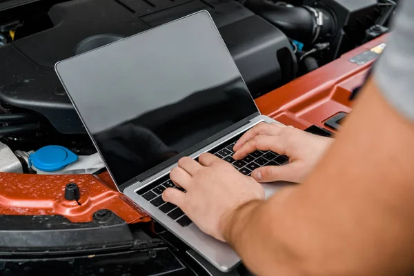 Partial view of auto mechanic working on laptop with blank screen at automobile with opened car cowl at mechanic shop — Stock Photo