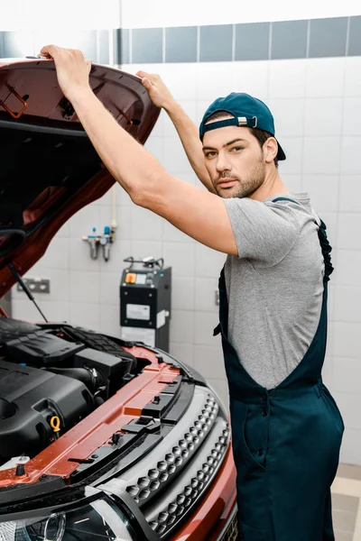 Automechaniker blickt in die Kamera, während er die Motorhaube in der Werkstatt untersucht — Stockfoto