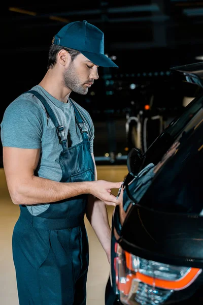 Vista lateral do automóvel jovem mecânico verificando o tronco do carro na oficina de reparação automóvel — Fotografia de Stock