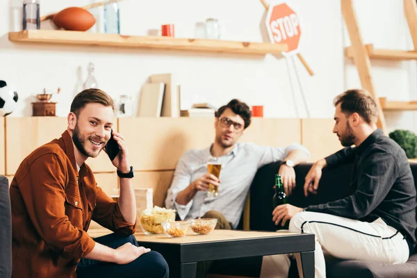 Enfoque selectivo de hombre sonriente con cerveza hablando en el teléfono inteligente con amigos detrás en la cafetería - foto de stock