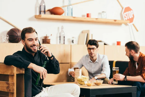 Enfoque selectivo de hombre sonriente con cerveza hablando en el teléfono inteligente con amigos detrás en la cafetería - foto de stock