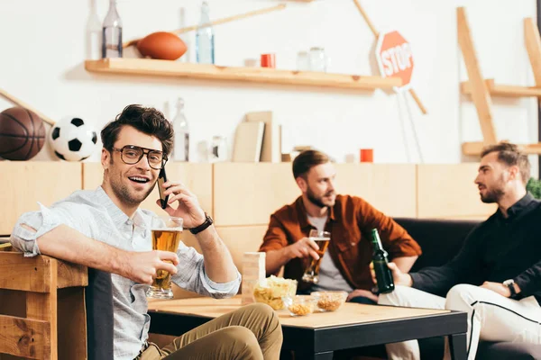 Selective focus of man with beer talking on smartphone with friends behind in cafe — Stock Photo