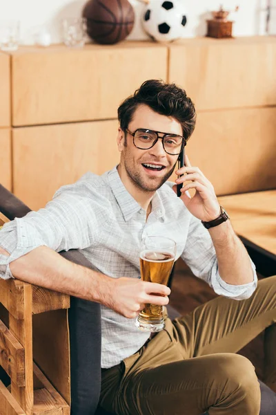 Портрет веселого чоловіка з келихом пива в руці розмовляє на смартфоні в кафе — стокове фото