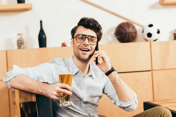 Портрет веселого человека со стаканом пива в руке разговаривающего по смартфону в кафе — стоковое фото