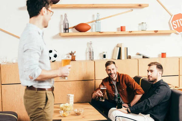 Hombre con cerveza mirando a los amigos descansando en el sofá en la cafetería - foto de stock