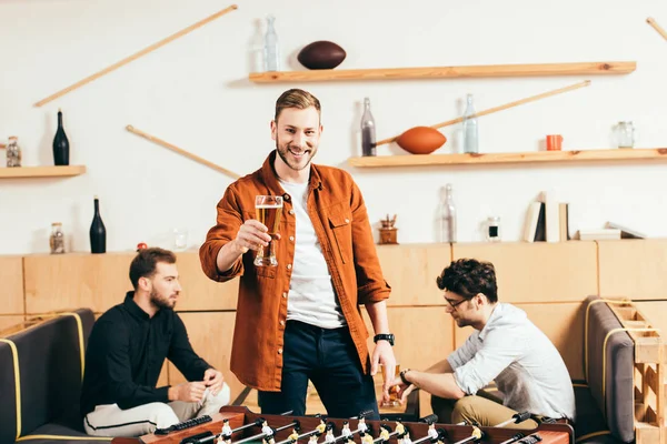 Вибірковий фокус усміхненого чоловіка зі склянкою пива, що стоїть на планшетному футболі в кафе з друзями позаду — стокове фото