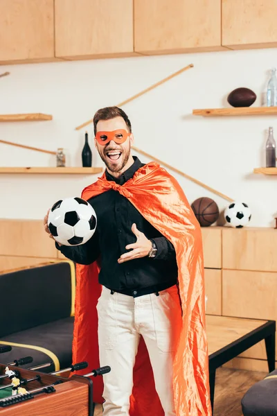 Joven emocionado en traje de superhéroe rojo con pelota de fútbol en la cafetería - foto de stock