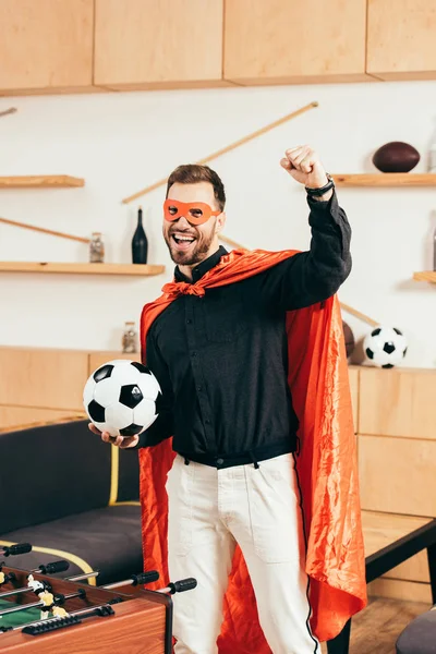 Happy young man in red superhero costume with soccer ball gesturing in cafe — Stock Photo