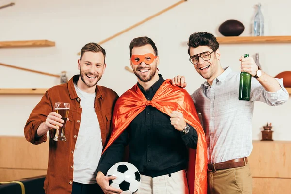 Портрет усміхненого чоловіка в костюмі супергероя з футбольним м'ячем в руці і друзями біля кафе — стокове фото