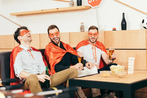 Cheerful friends in superhero costumes with beer resting together in cafe — Stock Photo