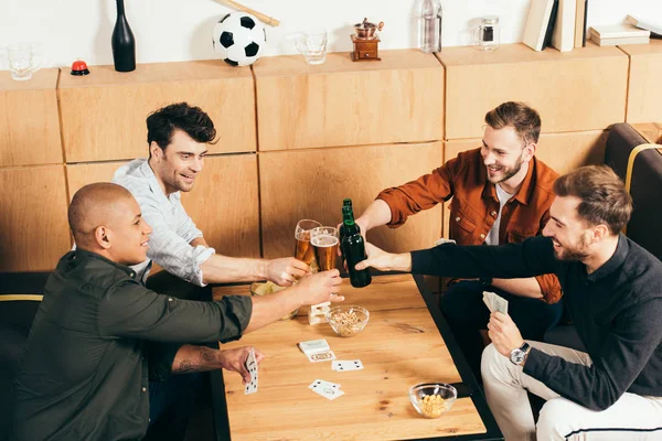 Amigos multirraciales sonrientes tintineando bebidas en la mesa con tarjetas y aperitivos en la cafetería - foto de stock
