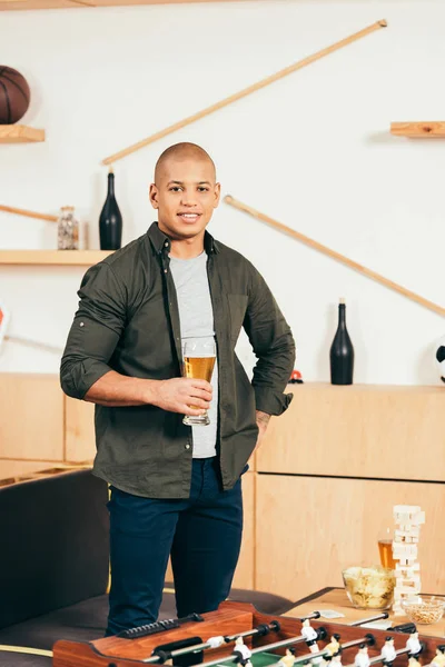 Smiling african american man with glass of beer standing in cafe with table soccer — Stock Photo