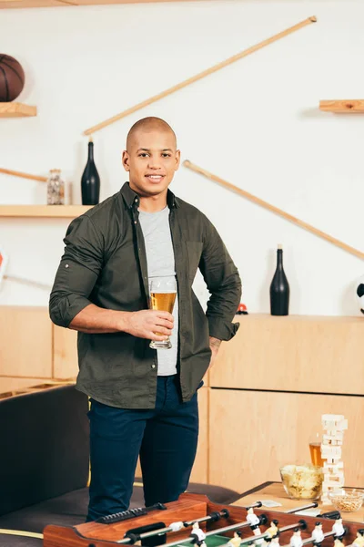 Sonriente afroamericano hombre con vaso de cerveza de pie en la cafetería con futbolín - foto de stock