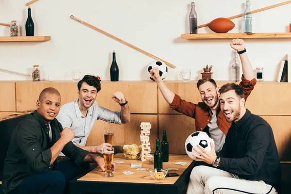 Amigos multiculturales felices haciendo gestos mientras se sienta en la mesa con cerveza y aperitivos en la cafetería - foto de stock