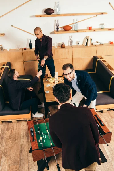 Vista de ángulo alto de los hombres de negocios jugando futbolín, mientras que los colegas multiétnicos tintineando bebidas detrás en la cafetería - foto de stock