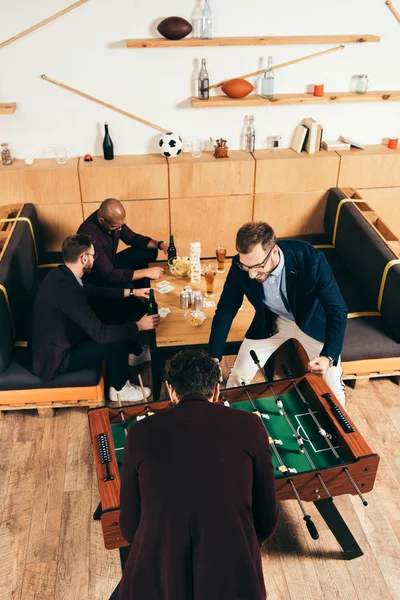 Vista de ángulo alto de los hombres de negocios jugando al futbolín mientras colegas multiétnicos descansan en el sofá en la cafetería - foto de stock