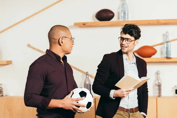 Retrato de empresarios multiétnicos con balón de fútbol y libro en la cafetería - foto de stock