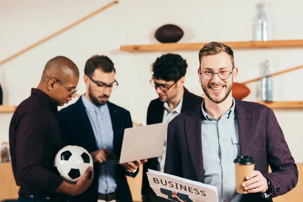 Enfoque selectivo de sonriente hombre de negocios con periódico y café para ir y colegas multiétnicos utilizando el ordenador portátil detrás en la cafetería - foto de stock