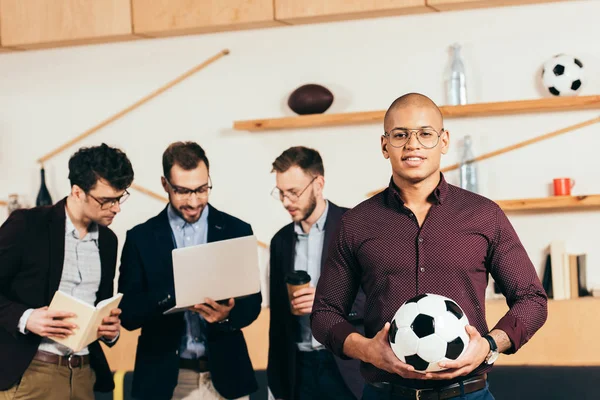 Enfoque selectivo de hombre de negocios afroamericano con pelota de fútbol y colegas multiculturales utilizando el ordenador portátil detrás en la cafetería - foto de stock