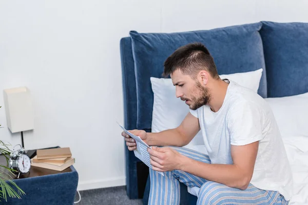 Side view of handsome man sitting on bed and reading newspaper in morning at home — Stock Photo