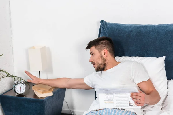Handsome man holding newspaper in bed and turning off alarm clock in morning at home — Stock Photo