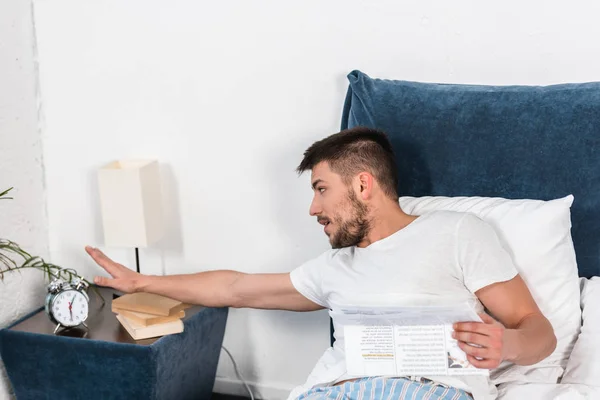 Hombre guapo sosteniendo el periódico en la cama y apagando el despertador en la mañana en casa - foto de stock