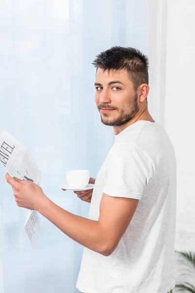 Hombre guapo leyendo el periódico de viaje y sosteniendo la taza de café en la mañana en casa - foto de stock