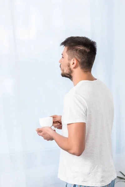 Vista lateral del hombre guapo de pie con taza de café en la mañana en el dormitorio - foto de stock