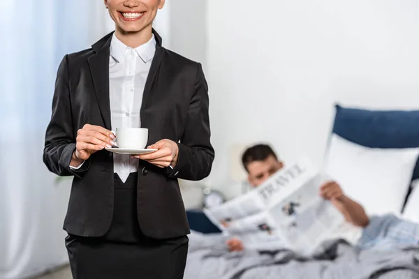 Cropped image of girlfriend in suit holding cup of coffee and boyfriend in pajamas reading newspaper in bedroom in morning, gender equality concept — Stock Photo