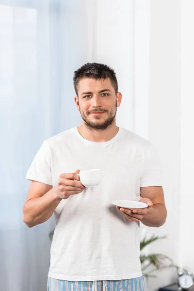 Красивий чоловік у сорочці і піжамі тримає чашку кави і дивиться на камеру вранці в спальні — стокове фото