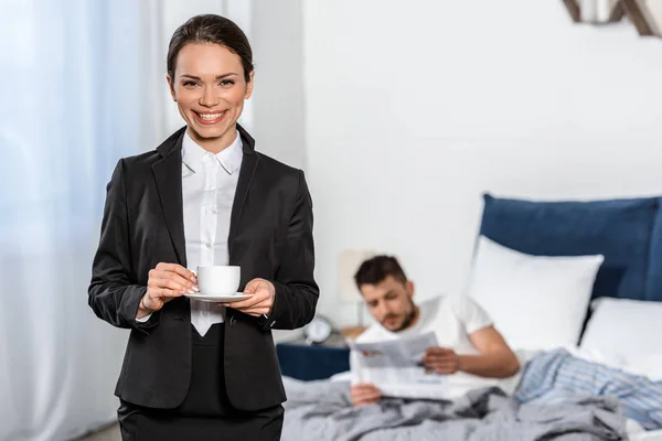 Petite amie souriante en costume tenant tasse de café et petit ami en pyjama lecture journal dans la chambre à coucher le matin, concept d'égalité des sexes — Photo de stock