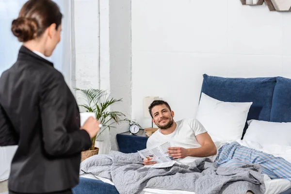 Girlfriend holding cup of coffee in suit and boyfriend in pajamas reading newspaper in bedroom in morning, feminist concept — Stock Photo
