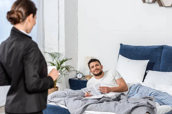 Petite amie tenant tasse de café en costume et petit ami en pyjama lecture journal dans la chambre le matin, concept féministe — Photo de stock